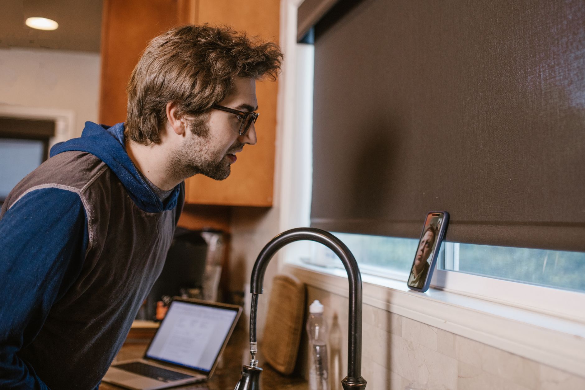 A man leans toward a phone propped by a kitchen window