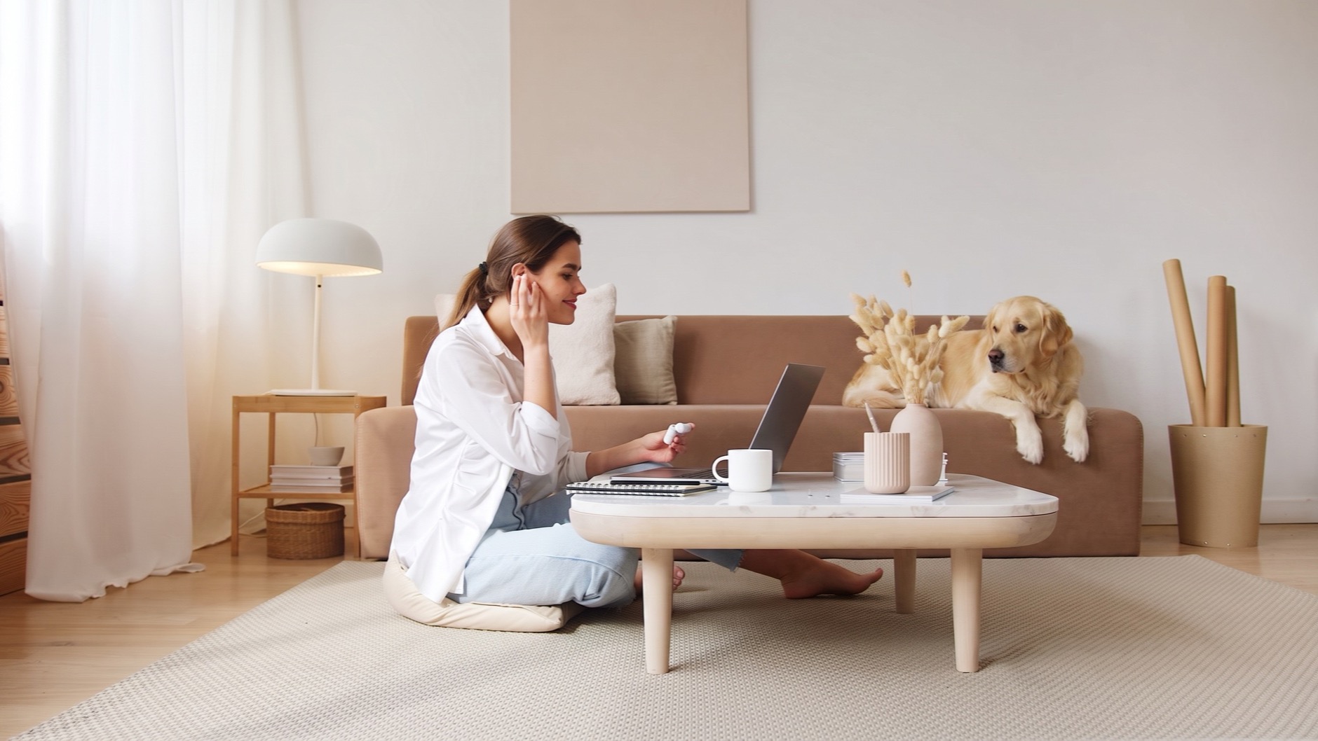 A woman with earbuds talks to a laptop from the living-room floor while her dog watches from the sofa